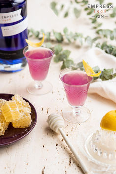 two glasses filled with pink lemonade next to a bottle of blueberry gin and a plate with slices of lemon on it