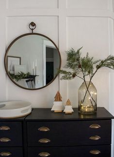 a black dresser topped with a vase filled with greenery next to a round mirror