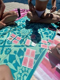 three people sitting on a towel playing cards by the water while one person sits in front of them