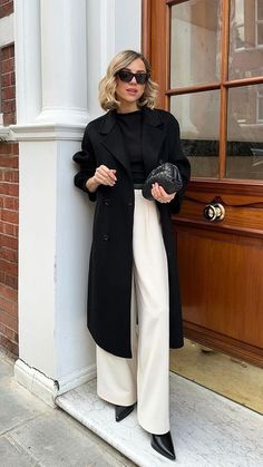 a woman in black coat and white pants leaning against a wall with her hand on her hip