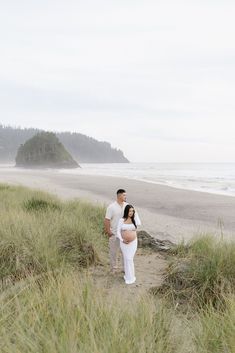 a pregnant couple standing on the beach in front of an ocean and sand dune area