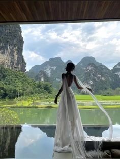 a woman in a wedding dress standing on a ledge looking out over a mountain lake