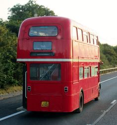 a red double decker bus driving down the road with trees in the backgroud