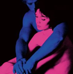 a man and woman sitting next to each other on a bed with pink and blue colors