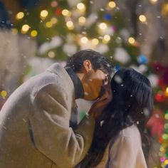 a man and woman kissing in front of a christmas tree with lights on the background