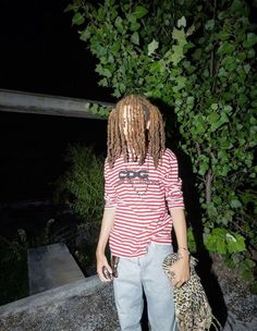 a man with dreadlocks standing next to a tree and holding a leopard bag