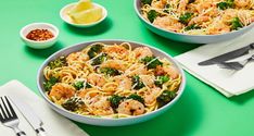 two bowls of shrimp and broccoli pasta on a green table with silverware