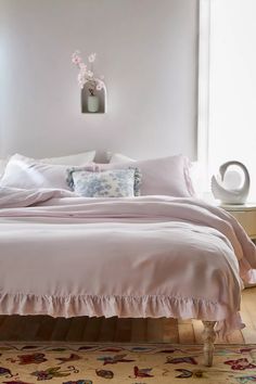 a bed with pink sheets and pillows in a white room next to a vase filled with flowers