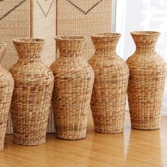 four vases are lined up on the floor in front of a wall with woven fabric