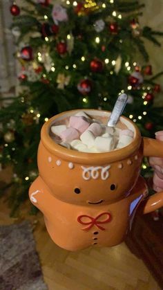 a person holding a mug with marshmallows in it near a christmas tree