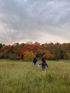 two people are walking through the grass in front of trees with orange and yellow leaves