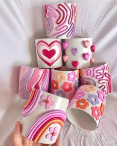 a hand is holding several cups with hearts and flowers painted on them, all in different colors