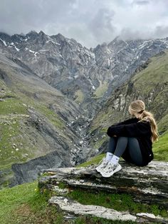 a woman sitting on the edge of a cliff looking at mountains with snow capped peaks in the distance