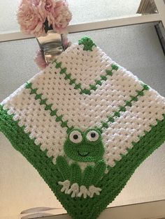 a crocheted pot holder with a frog on it's side and flowers in the background