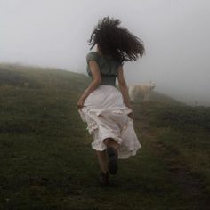 a woman in a white dress walking down a foggy hill with a dog behind her