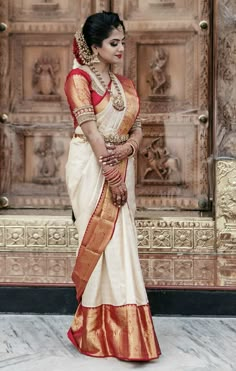 a woman in a white and red sari with gold trims standing by a wooden door