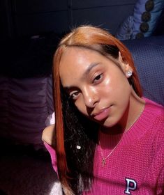 a woman with long red hair wearing a pink sweater and pearl earrings sitting on a couch