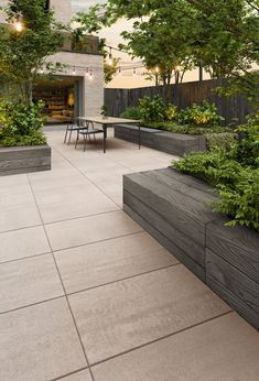 an outdoor dining area with tables and benches surrounded by trees, shrubs and plants on the side walk