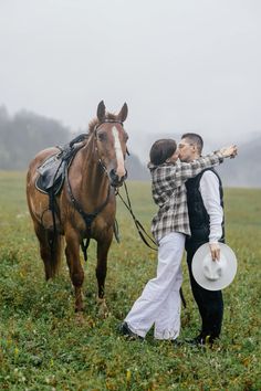 two people standing next to a brown horse on a lush green field with foggy sky in the background