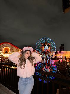 a woman in pink jacket standing next to ferris wheel and carnival rides at night time