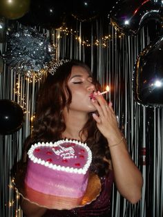 a woman is blowing out the candles on her heart - shaped cake in front of balloons and streamers