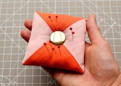 a hand is holding an orange and pink pin cushion with buttons on the front, while another button has been sewn onto it