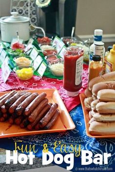 hot dogs and condiments sit on plates at a table with an advertisement for the hot dog bar