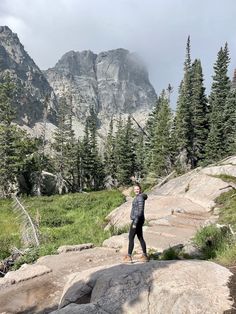 a woman standing on top of a rock in front of some trees and mountain peaks