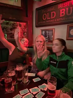 three women sitting at a table with beer glasses in front of them and one woman raising her hand