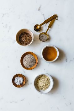 four bowls filled with different types of spices on top of a white counter next to two wooden spoons