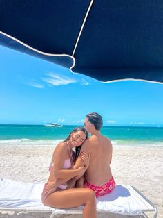 a man and woman sitting on the beach under an umbrella, cuddling up to each other