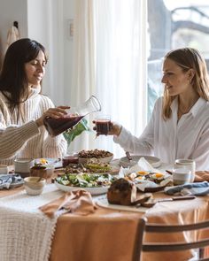 two women sitting at a table with food and drinks in their hands, one is pouring something into the other