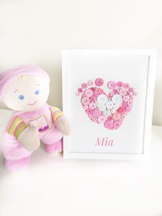 a pink teddy bear next to a framed personalized button heart art piece on a white surface