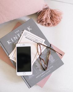 an iphone, eyeglasses, and book on a bed with a pink pillow