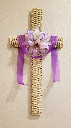 a cross made out of wicker with purple ribbon and flowers on it hanging from the wall