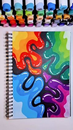 a spiral notebook with watercolors on it next to some pens and paper clips