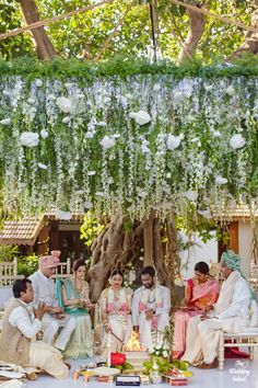 a group of people sitting around each other under a tree with flowers hanging from it