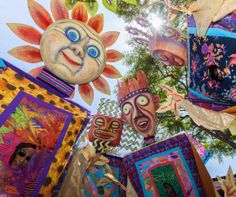 many colorful masks hanging from the ceiling in front of trees and blue sky with sun shining through them