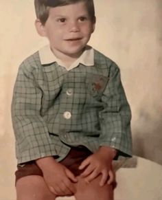 an old photo of a young boy sitting on top of a white chair with his legs crossed