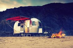 a camper trailer is set up next to a fire on the beach with mountains in the background