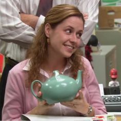 a woman sitting at a desk with a teapot in her hand and another man standing behind her