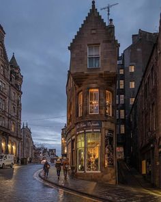 two people walking down the street in front of some buildings at night with lights on