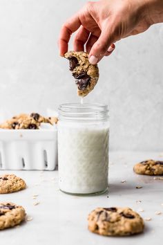 a hand holding a chocolate chip cookie over a jar of milk and cookies on the table