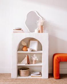 a white shelf with books, vases and other items on it next to an orange chair