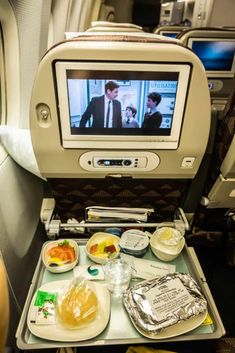 an airplane seat with food and drinks on the tray in front of it, while someone is watching tv