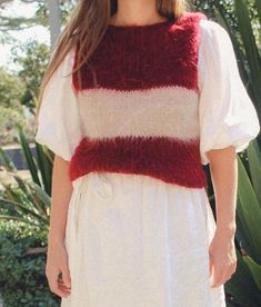 a woman wearing a white dress with red and white stripes on the chest, standing in front of some plants