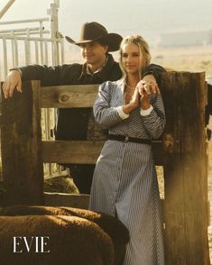 a man and woman standing next to a wooden fence with sheep in the back ground