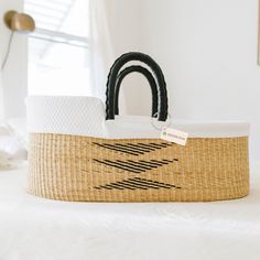 a woven basket with black handles sits on a white bed in front of a window