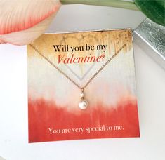 a card with a heart and a pearl on it next to a pink flower that says, will you be my valentine?