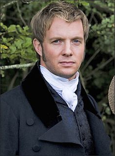 a man with blonde hair wearing a black coat and white shirt in front of trees